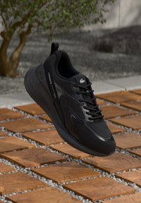 Černá atletická obuv s texturovaným svrškem, šněrováním a výrazným podpatkem, vystavená nad vzorovanou kamennou cestou. Přítomné logo Lacoste.