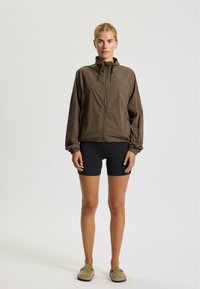 Lichte bruin jas met een aansluitende capuchon, gathered manchetten en een rits aan de voorkant; gestyled met zwarte shorts en beige instap schoenen.