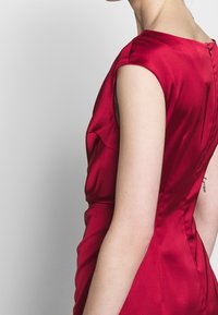Robe en satin rouge à la silhouette ajustée, manches courtes et fermeture éclair à l'arrière. Caractérisée par une texture lisse et des détails subtils de drapé.