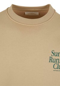 Hanorac bej cu guler ribfrat, având inscripții verzi "Sun Run Club" și "ANOTHER COTTON" imprimate pe față. Material moale, texturat.