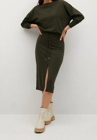 Frau trägt ein olivgrünes Langarmkleid mit einem Schlitz vorne und beigefarbenen Stiefeletten, steht vor einem schlichten weißen Hintergrund.