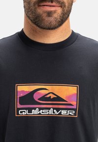 Sort t-shirt med korte ærmer, der har et farverigt Quiksilver-logo. Logoet viser en bølge og et bjerg i nuancer af lilla og orange.