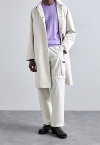 Ljusbeige regnjacka med svarta knappar, kombinerad med en lavendelfärgad tröja och krämfärgade byxor. Svarta ankelskotsar fulländar outfiten.