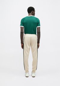 Πράσινο T-shirt με κοντά μανίκια και λευκή λεπτομέρεια, σε συνδυασμό με μπεζ φόρμες με μαύρες ρίγες στο πλάι. Το μοντέλο εμφανίζεται από πίσω.