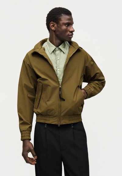 Férfi olíva zöld cipzáras dzsekit visel világos zöld gombos ing és fekete nadrág fölött, jobbra néz, bal kezét zsebre dugja.