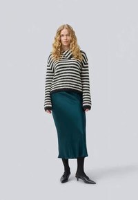 Pull en tricot rayé noir et blanc avec un décolleté large, associé à une jupe midi en satin teal et des collants noirs. Le mannequin se tient devant un fond neutre.