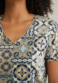 V-veida T-krekls ar izstrādājumu zīmējumu zilā, oranžā un zaļā krāsā, izgatavots no mīksta auduma ar gludu tekstūru.