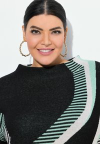 Femme aux cheveux bruns, coiffée de manière lisse, portant de grandes boucles d'oreilles en or et un haut noir à motifs rayés menthe et blanc, souriant à la caméra.