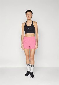 Mujer de pie, vistiendo un sujetador deportivo negro, pantalones cortos rosas, medias altas blancas con rayas negras y zapatillas deportivas negras contra un fondo blanco.