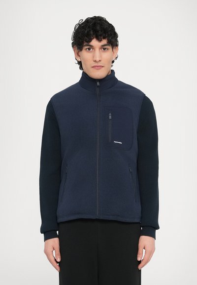 Veste sans manches en polaire bleu marine avec un col montant, une fermeture éclair frontale et des manches en maille. Comprend une poche poitrine et deux poches latérales.