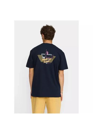 Hombre con camiseta azul marino con gráfico de "Pray for Surf" e ilustración de surfista en la espalda, combinado con pantalones beige.