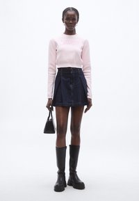 Maglione corto rosa chiaro con logo "tommy jeans", abbinato a una gonna a linea A in denim scuro. La modella tiene in mano una piccola borsa nera e indossa stivali neri al ginocchio.