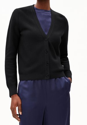 Vrouw die een zwart gilet met knopen draagt over een marineblauwe blouse en bijpassende marineblauwe broek, staand met één hand in de zak.