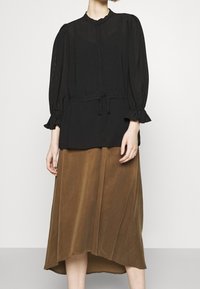 Blouse noire texturée avec un col rond et un cordon de serrage, associée à une longue jupe brune avec un ourlet asymétrique.