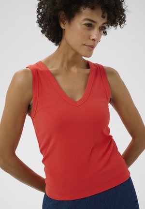 Vrouw met krullend haar, gekleed in een mouwloze rode V-hals geribbelde top en donkerblauwe broek, poseert met haar handen achter haar rug tegen een effen achtergrond.