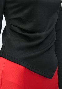 Μαύρο μπλουζάκι με μακριά μανίκια και λεία υφή, με ασύμμετρο τελείωμα. Συνδυάζεται με φωτεινή κόκκινη φούστα, δημιουργώντας έντονη αντίθεση χρωμάτων.