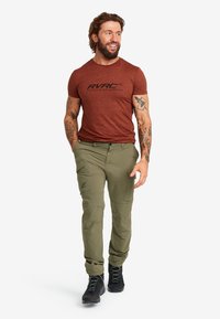 Gröna cargobyxor med fickor och en brun kortärmad t-shirt med en logotyp. Bärs med svarta vandringsskor. Modellen ler och går.