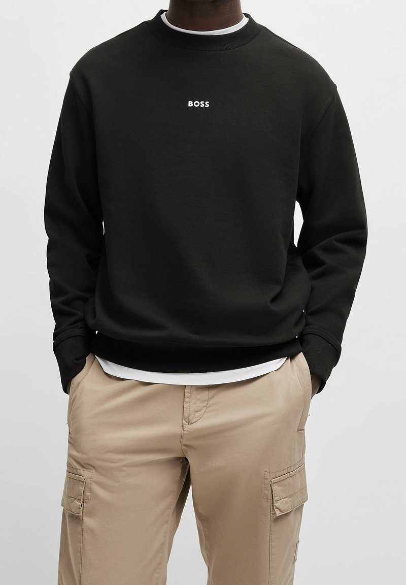 Homme portant un sweat-shirt noir avec le mot "BOSS" centré sur la poitrine, un pantalon cargo beige et les mains dans les poches, sur un fond uni.