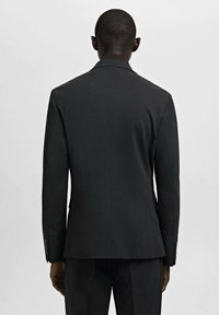 Man staat met zijn gezicht van de camera af, gekleed in een nauwsluitend zwart colbert en bijpassende broek, voor een effen lichte achtergrond.