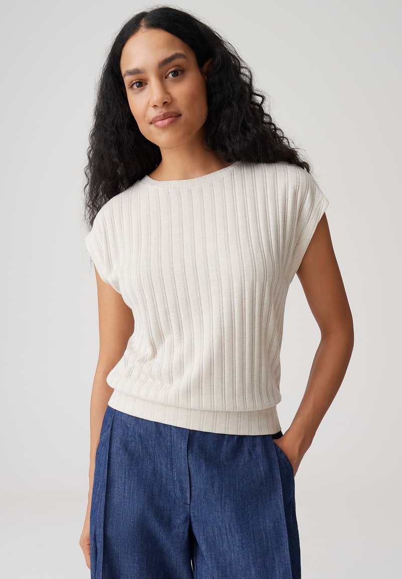 Donna con lunghi capelli ricci neri, che indossa un top bianco smanicato a coste e pantaloni blu larghi, in piedi con una mano in tasca.