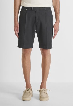 Hombre con pantalones cortos negros hasta la rodilla y zapatos casuales beige con cordones sobre fondo liso.