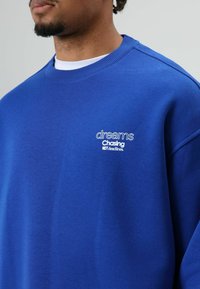 Sweat-shirt bleu à encolure ronde, avec un texte brodé blanc. Le tissu semble doux avec une texture lisse.