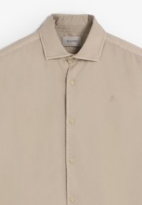 Camisa beige de botones confeccionada en una tela suave. Presenta un cuello puntiagudo, cinco botones y un pequeño logo bordado en el lado izquierdo.