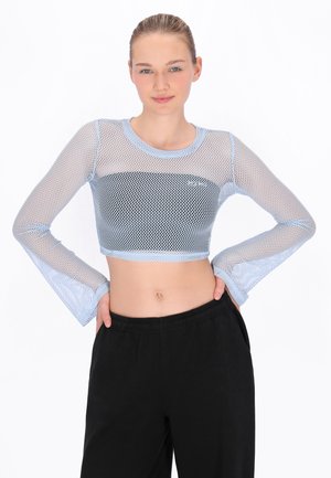 Junge Frau mit zurückgebundenem Haar, die ein hellblaues Netz-Crop-Top mit langen Ärmeln und schwarze Hosen trägt, Hände auf den Hüften, neutraler Hintergrund.