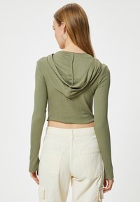 Sudadera corta de manga larga en tela de canalé verde oliva; cuenta con una capucha drapeada y varios detalles de costura; combinada con pantalones cargo de color claro.