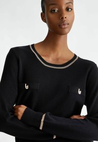 Maglione nero tessuto con collo rotondo, dettagli in argento e due tasche frontali con ferramenta decorativa. Maniche lunghe con bordi argentati.