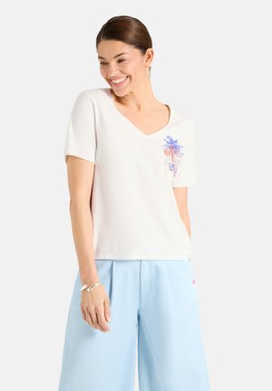 Frau lächelt und schaut nach unten, trägt ein weißes V-Ausschnitt-Top mit floraler Stickerei und hellblaue, weit geschnittene Hosen.