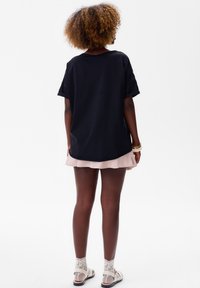 Zwarte katoenen t-shirt met korte mouwen, ontspannen pasvorm en ronde halslijn, gecombineerd met een lichtroze plissé rok en witte sandalen.