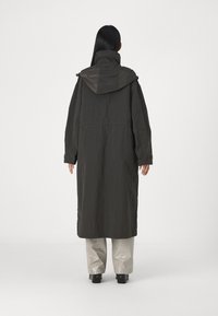 Parka longue de couleur vert foncé avec capuche, présentant une coupe ample et des manches à poignets. Le matériau semble texturé avec une finition mate.