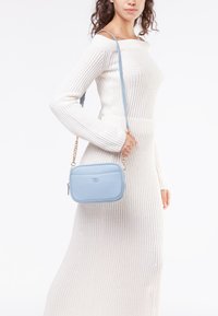 Borsa a tracolla blu chiaro con finitura testurizzata, forma rettangolare e dettagli in catena dorata, indossata da un modello su uno sfondo neutro.