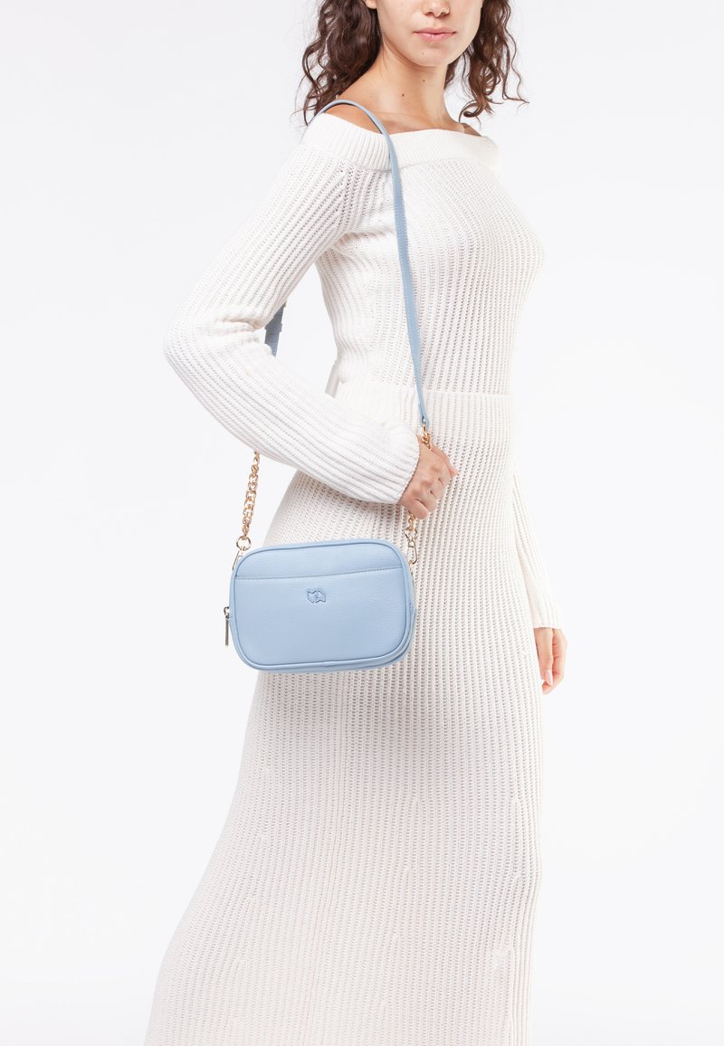 Borsa a tracolla blu chiaro con finitura testurizzata, forma rettangolare e dettagli in catena dorata, indossata da un modello su uno sfondo neutro.