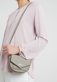 Harmaa nahkainen olkalaukku, jossa on kolmionmuotoinen läppä ja tikattu koristelu, kantaa henkilö, joka pukeutunut vaaleanpunaiseen puffhihalliseen paitaan.