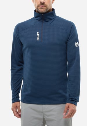 Homme portant une polaire demi-fermeture éclair bleu marine texturée avec le logo "Millet" sur la poitrine et la manche, associée à un pantalon gris clair.