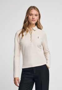 Koszulka polo z długim rękawem w jasnobeżowym kolorze, z beżowym kołnierzykiem i pięcioma guzikami. Na lewej piersi znajduje się małe, brązowe, haftowane logo.