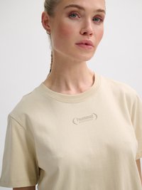 Frau mit geflochtenem Haar, die ein beiges hummel Sport-T-Shirt mit kleinem Logo auf der Brust vor einfachem Hintergrund trägt.