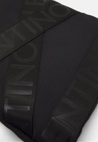 Svart texturerad tygaccessoar med en diaganlrem och präglad logotyp. Designen inkluderar en mönstrad yta och en strömlinjeformad form.