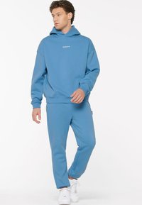 Blaues Kapuzenshirt und passende Hose aus weichem Stoff. Mit einer Kängurutasche und gerippten Bündchen. Getragen mit weißen Turnschuhen.