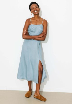 Femme souriante aux cheveux courts et bouclés portant une robe midi sans manches bleu clair avec une fente latérale et des chaussures marron à enfiler, debout les bras croisés.