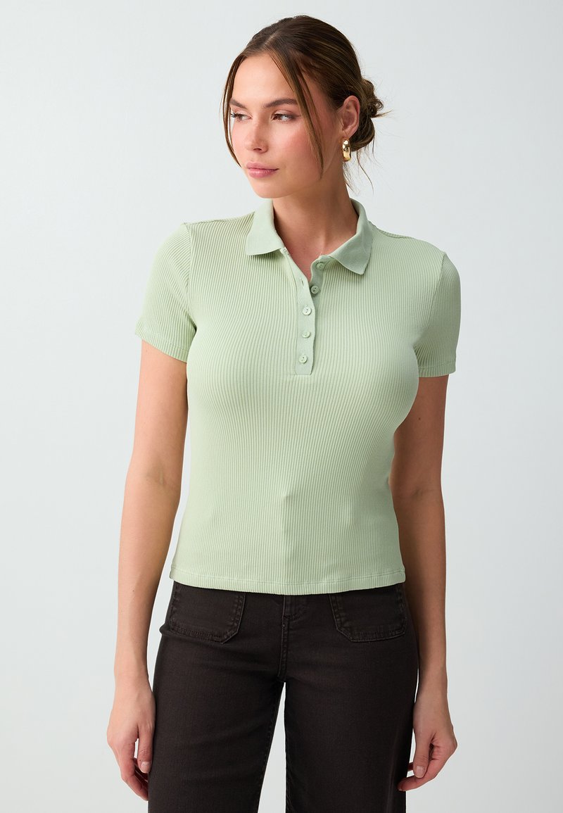 Femme avec des boucles d'oreilles créoles dorées, portant un polo côtelé à manches courtes vert clair et un pantalon noir, regardant sur le côté sur un fond uni.
