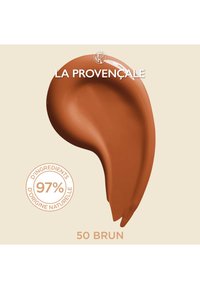 Produit cosmétique marron en forme de coup de pinceau, étiqueté "50 Brun" avec une note sur 97% d'ingrédients naturels, posé sur un fond neutre.