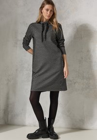 Mekko, jossa on musta ja harmaa kuvio, pitkät hihat, solmittava kaula-aukko, rento malli ja suora leikkaus, yhdistetty mustiin paksupohjaisiin saappaisiin.