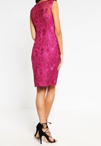 Fuchsiové krajkové šaty s přiléhavým střihem, bez rukávů a složitými květinovými vzory. Vyznačují se černými sandály na vysokém podpatku s řemínky.