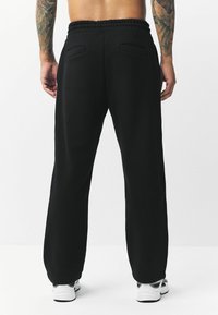 Schwarze Sweatpants mit einem elastischen Bund, zwei Gesäßtaschen und einer lockeren Passform. Weiche Textur, kombiniert mit weißen Sportschuhen.