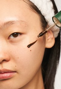 Un siero marrone viene applicato con un contagocce sulla guancia di una persona che indossa una fascia bianca e nessun trucco.