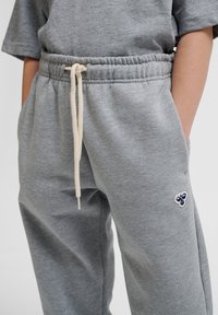 Graue Jogginghosen mit einem elastischen Bund und beigem Kordelzug. Sie verfügen über seitliche Taschen und ein kleines blaues gesticktes Design am Bein.