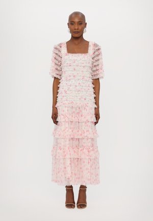 Robe à volants blanche et rose en tissu transparent avec des détails froncés. Encolure carrée et manches bouffantes. Sandales à talons brunes.